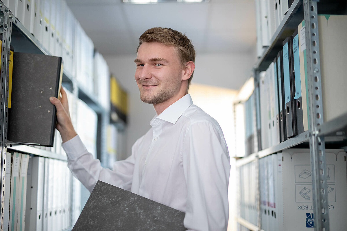 Ein BIT Kollege im Standort Karlsruhe Ein Kollege mit Ordnern in der Hand steht vor einem Regal.